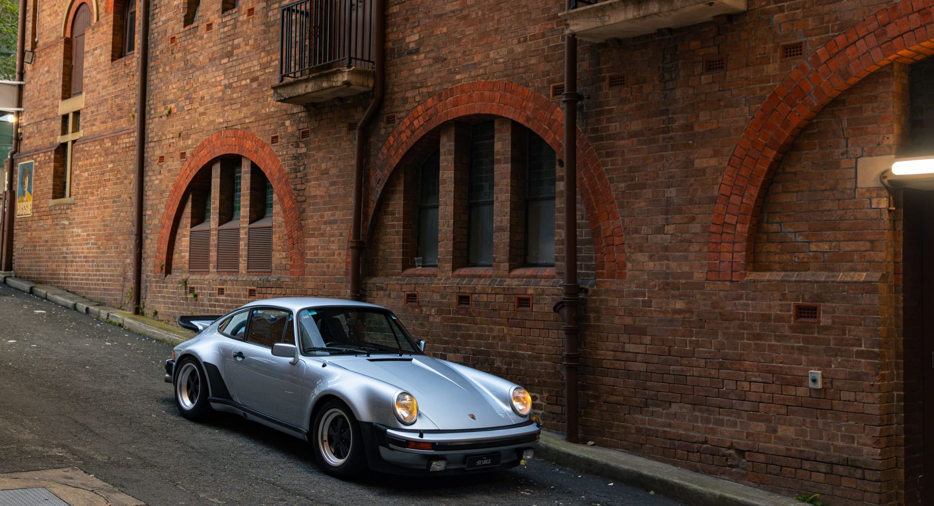 Silver air-cooled Porsche 930 Turbo beside the red brick arches of a Brisbane warehouse