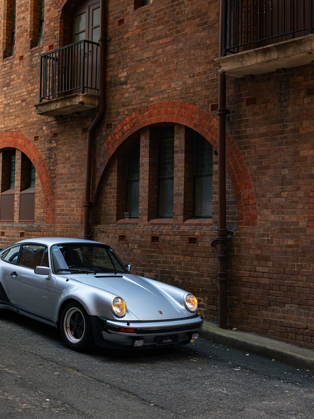 Silver air-cooled Porsche 930 Turbo beside the red brick arches of a Brisbane warehouse